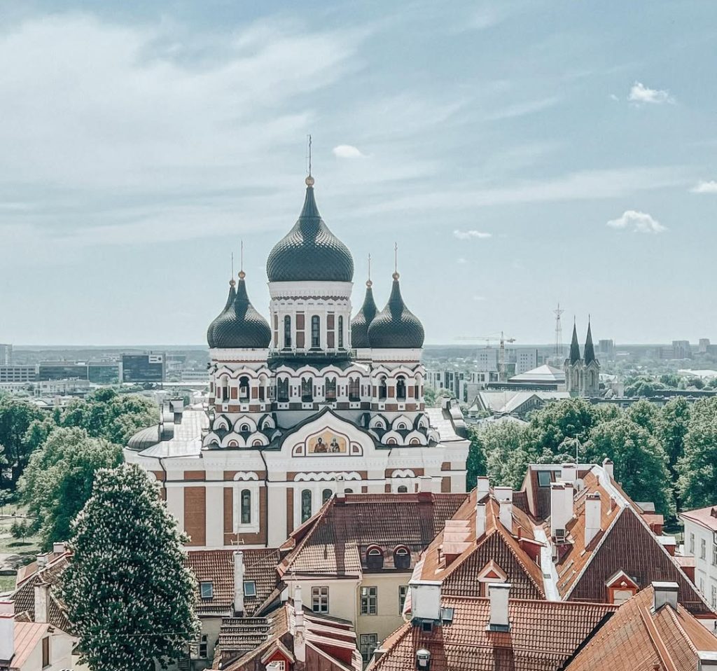 A scenic view of the Alexander Nevsky Cathedral in Tallinn, Estonia, with its distinctive black domes rising above the surrounding rooftops and greenery on a clear day.