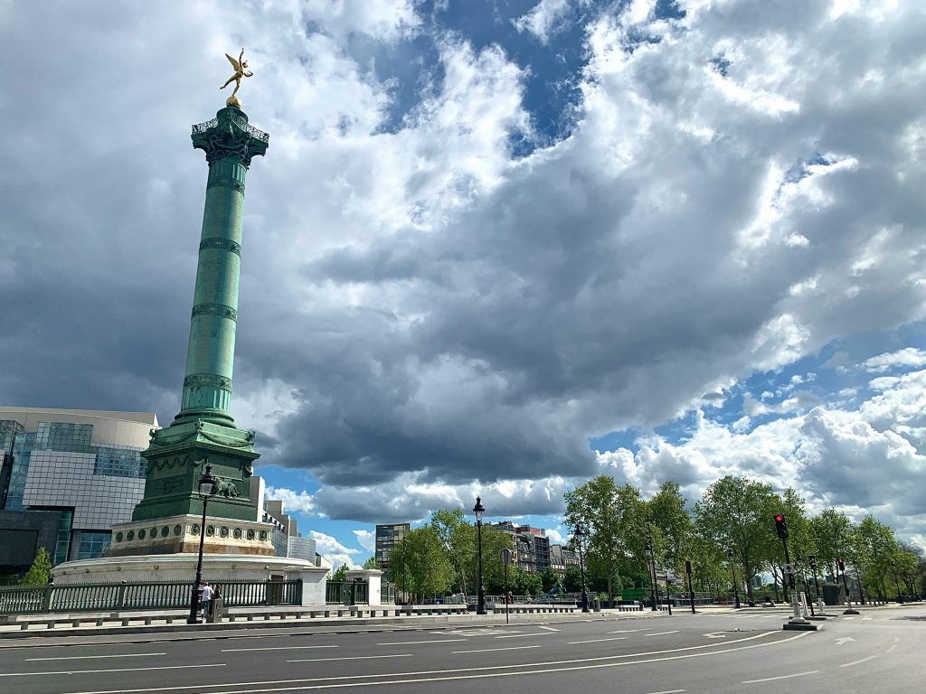 A striking view of the Bastille Monument in Paris, with the towering green column and its golden statue set against a dramatic cloudy sky.