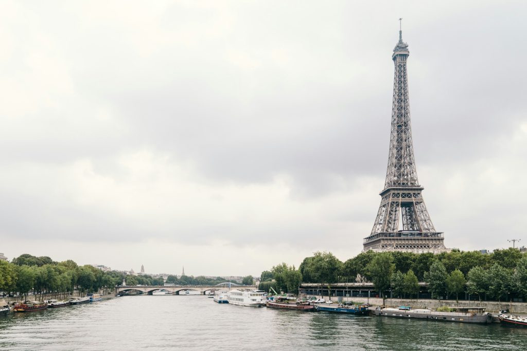 A serene view of the Eiffel Tower, Paris, rising above the Seine River with boats along the riverbank and a cloudy sky overhead.