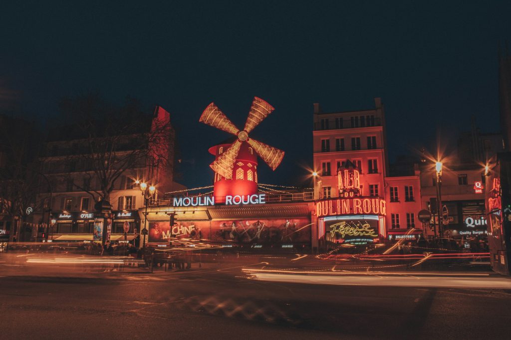 A vibrant night scene of the iconic Moulin Rouge in Pigalle, Paris, with its famous red windmill glowing brightly against the dark sky.